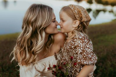 An adult woman and a young girl, both smiling and touching noses, sit outdoors by a lake during a Warner Robins family photography session at Flat Creek, beautifully captured by Fire Family Photography. The girl holds flowers and wears a patterned headscarf and dress. 
