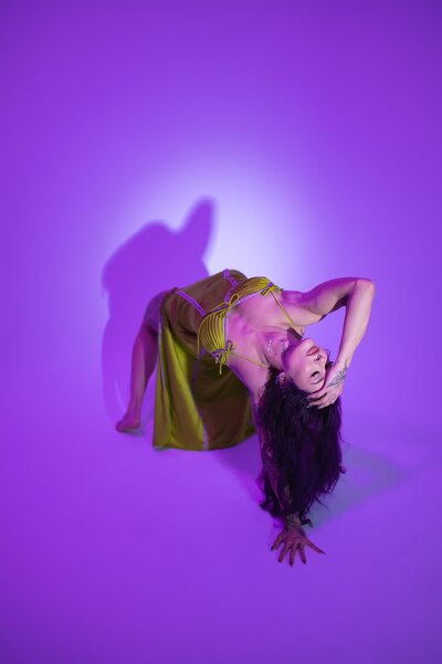 Woman with long, black hair is in a backbend with one arm reaching over her head as she closes her eyes wearing a silky green dress in front of a purple backdrop.