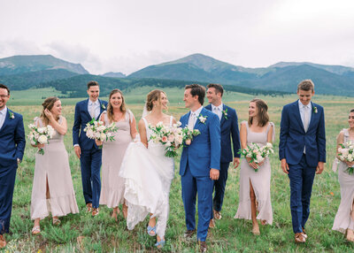 A bridal party dressed in blues and pastel pinks walk and and laugh together during portraits while the bride and groom walk arm in arm
