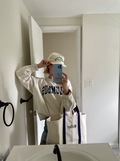 Young Woman taking mirror selfie wearing a baseball cap and holding tote bag