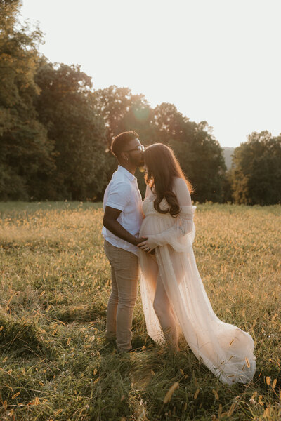 Couple kisses in field while holding the pregnant belly for their golden hour portrait session in Coopersburg PA 