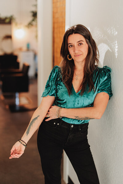 Chelsea a hair stylist in Eugene leans against a wall while holding out her arm featuring a feather tatoo.