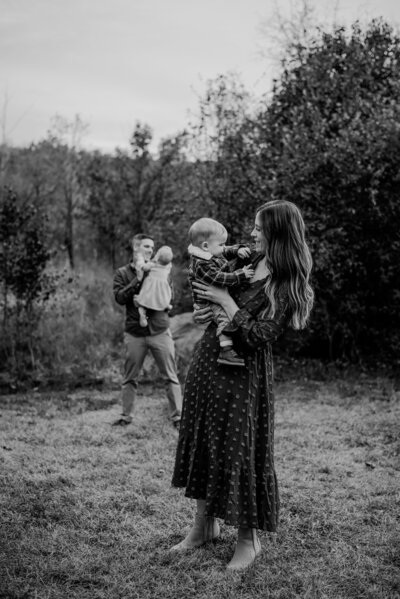 Black and white photo of mom holding baby standing close to the camera and dad holding their other baby behind mom.