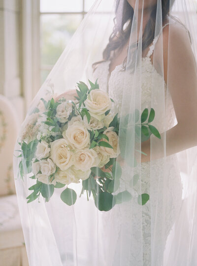 Close-up of a bride holding a lush white and green wedding bouquet, captured in soft, natural light