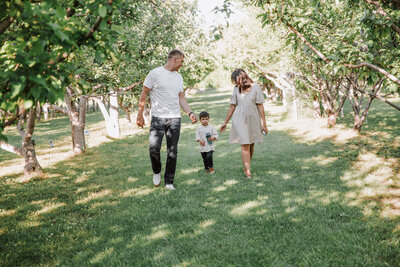 Idaho Falls family photographer captures family of three holding hands and walking down a path in an orchard in the summer time