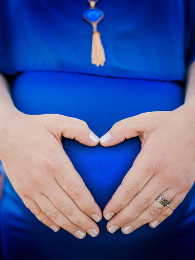 A pregnant mother in a bright blue dress and blue-gold necklace holds her hands in the shape of a heart over her belly. Photo by SAVI Photography - San Diego California Photographer