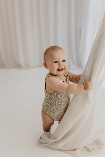 A little baby boy holds onto the curtain and giggles during a action photo in a milestone photoshoot with lucy jane photography in christchurch