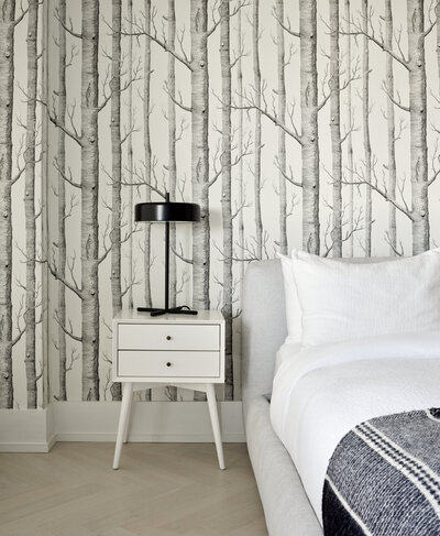 A small white side table with two drawers holding a black lamp next to a grey and white bed with monochrome tree artwork on the wall behind.