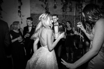 Bride is holding a drink smiling and dancing .  Groom is smiling and dancing.  Guests are standing watching bride and groom dance in the London wedding venue