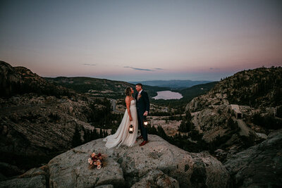 A bride and groom holding lanterns gaze lovingly at each other while standing atop a rocky viewpoint near Lake Tahoe at sunset, with Donner Lake and a serene mountainous landscape visible in the distance.