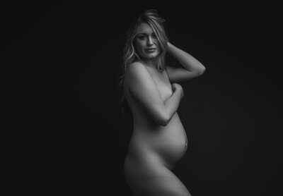 Black and white tasteful nude maternity portrait with pregnant mother looking into the camera will pushing her hair up