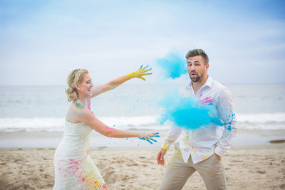 Married couple having fun on the beach with colors in a paint war as they trash the dress
