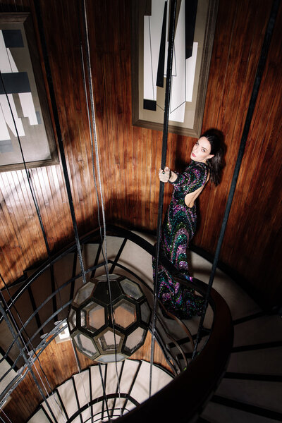 Fashion portrait of model in long evening gown standing on spiral staircase with flash lighting