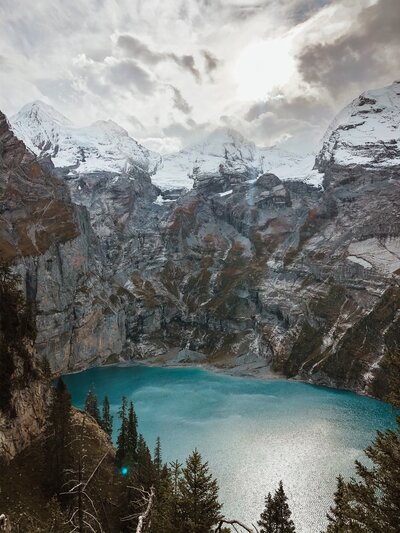 Alps in Switzerland. They are snow capped with a bright blue lake right below. There are pine trees coming up from the alps and rocks surrounding the base of the lake.