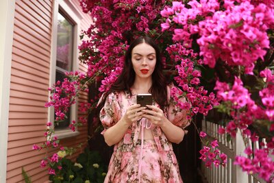 Woman in a pink dress surrounded by pink flowers looking at her phone. Photo by Hiki App on Unsplash