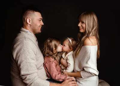 family of 4 on black background and siblings kissing so sweetly and mom and dad smiles at one another