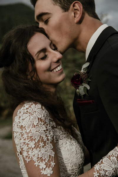 close up image of a bride and groom at rolley lake. The bride's hand is on the grooms arm and he is leaning in to kiss her temple. They both have their eyes closed and the bride is smiling.