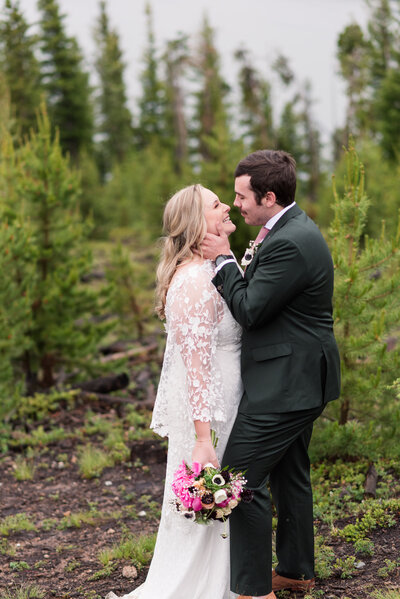 Rocky Mountain National Park wedding with bride and groom embraicng one another in a forrest as the groom caresses the brides cheek while she holds her pink floral bouquet to her side photographed by denver wedding photographer