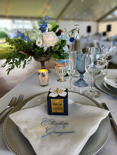 A Tablesetting including a table with a napkin with blue printed names, blue and white flowers, candles, and blue and white beverage glasses