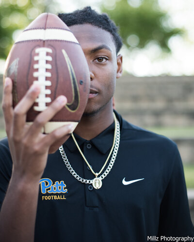a senior posing for graduation photos holding a football in front of his face photographed by Millz Photography in Greenville, SC