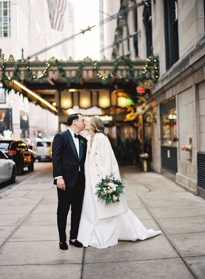 Bride and groom share a kiss on a city street, with the bride in a long white gown and a fur coat, holding a large green and white floral bouquet. Festive garlands and lights adorn the building facade behind them, adding a romantic winter holiday atmosphere to the scene.