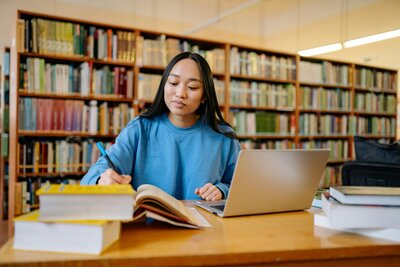 A woman studies at a library table with books and a laptop, inspired by The Summer Jumpstart with Mya Andersen to get college-ready this summer.