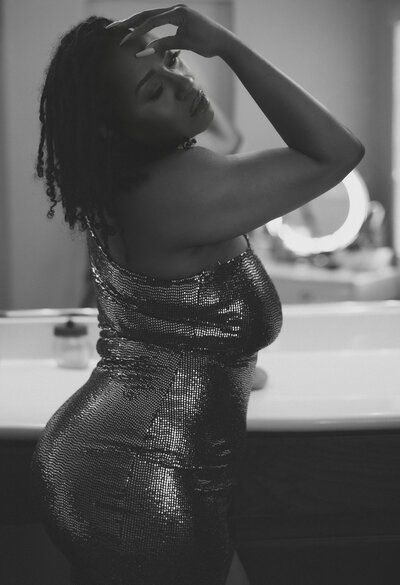 lack and white boudoir portrait of a confident woman in a shimmering dress, softly lit as she poses in front of a bathroom mirror with her hand resting on her forehead. Her expression is calm and introspective, capturing a quiet moment of sensuality and self-reflection.