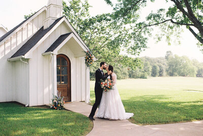 A newlywed couple, dressed in wedding attire, shares an intimate kiss outside a charming white chapel, surrounded by lush green grass and trees under a bright sky. The bride holds a vibrant bouquet of flowers.