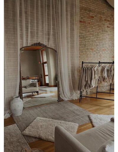 A large mirror leaning up against a brick wall and a clothing rack standing next to it with neutral colored clothing hanging.