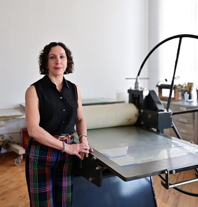 Printmaker Nichol Markowitz in a black and white dotted blouse leans on a large rolling press machine in a light-filled printmaking studio.