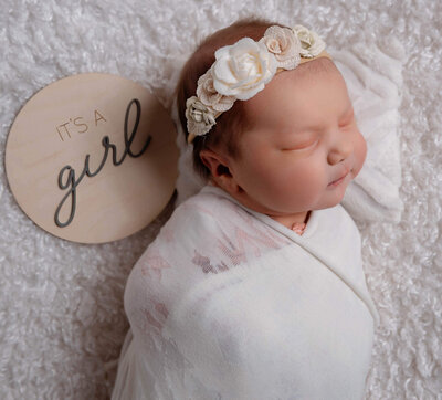 newborn baby girl with floral head bow and wooden sign that says its a girl
