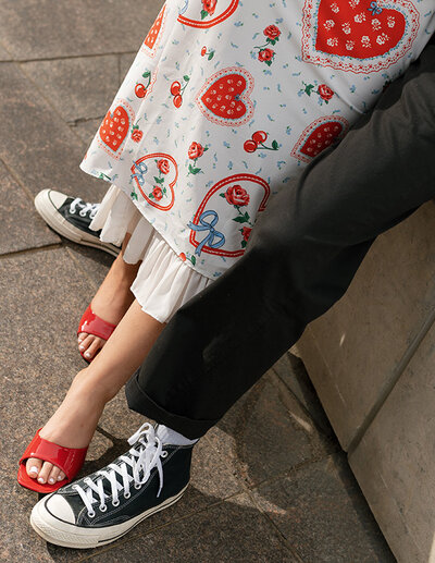 A person wearing a heart-patterned dress and red sandals stands beside someone in black pants and sneakers. The scene conveys a casual, playful vibe.