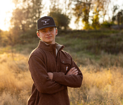 golden hour senior boy photos - he is wearing a black hat and rust colored sweater with his arms crossed - in a field at sunset in the pacific northwest