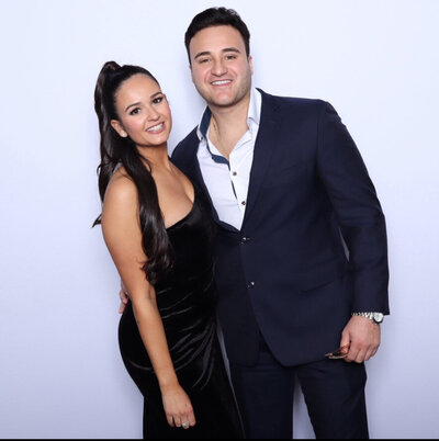 A couple dressed in formal attire posing closely in front of a white backdrop, with the woman in a black dress and the man in a navy suit.