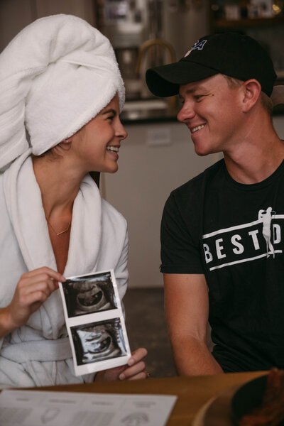 Woman in a robe and towel hair wrap holds a sonogram images and smiles at her partner.