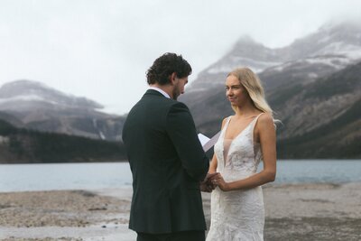 bride smiles at groom as he reads vows on a beach with mountains in background in Banff