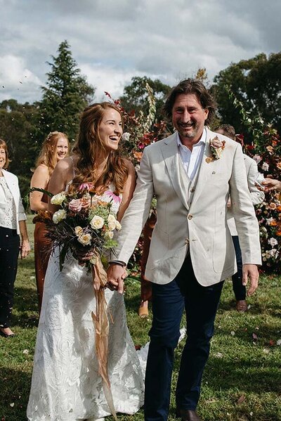 Bride holding a bouquet and walking hand-in-hand with the groom down a grassy aisle, surrounded by cheering guests at an outdoor Brisbane wedding.