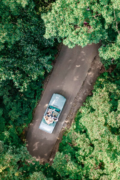Aerial view of an open-top vehicle driving along a narrow, paved road through lush green trees. The road is surrounded by dense forest, and a group of people can be seen inside the vehicle.