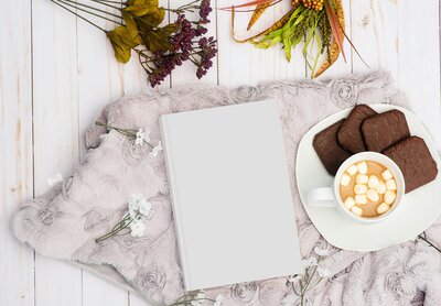 Un livre à la couverture blanche posé sur un plaid à côté d'une tasse avec un café et des petits gâteaux