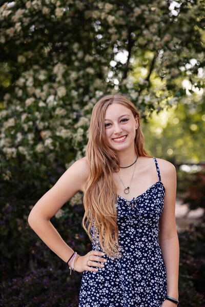 Girl in blue dress and long blonde hair puts hand on hip and smiles for her senior photos.