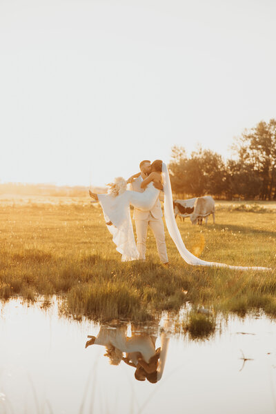 Groom lifting bride for a kiss beside a pond during golden hour at their Montana wedding.