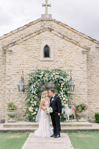 Bride and groom standing at stone chapel with lush floral arch backdrop
