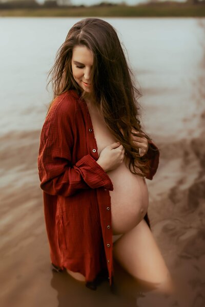 A pregnant person stands knee-deep in the waters of Flat Creek Perry GA, wearing an open, oversized red shirt with long sleeves. They have long, wavy hair and are touching their shirt with one hand while looking down, capturing a serene moment perfect for fire family photography.