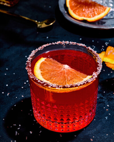 A striking close-up of a red cocktail garnished with a citrus slice and a salt rim, captured by Austin-based food photographer Mica McCook. The vibrant colors and detailed texture highlight the artistry in mixology photography.