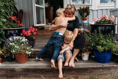 Candid family moment on a flower-filled porch—grandmother sitting barefoot while embracing her two young grandchildren in a warm, emotional hug.