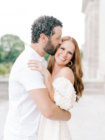 Curly-haired husband kisses redheaded wife's cheek while she giggles at the camera