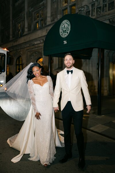 groom holds bride's dress train as they walk down a path