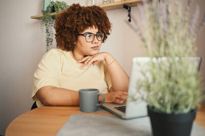 A black woman assistant professor gazes at her laptop with a neutral expression. There is a coffee mug beside her computer and plants in the foreground and background.