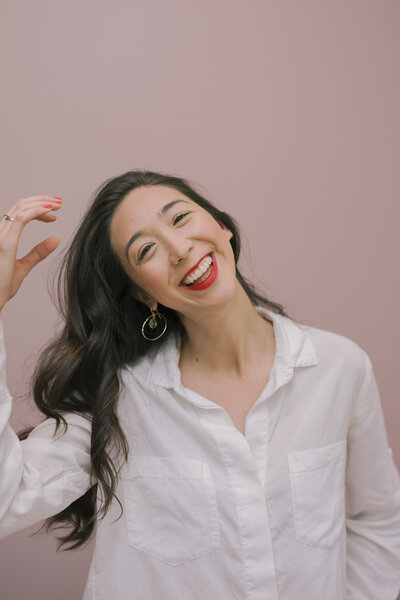 A woman smiling with red lipstick and posing in front of a pink wall while playing with her hair