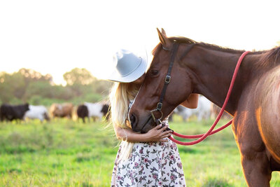 girl wearing cowboy hat while kissing her horse's nose with cows in the background